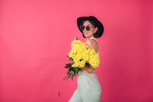 Beautiful stylish woman in hat and sunglasses posing over pink red background holding large bouquet of yellow asters, spring mood, positive emotions isolated space - Photo, image