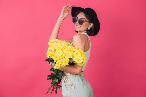 Bella donna elegante in cappello e occhiali da sole in posa su sfondo rosso rosa con grande bouquet di astri gialli, umore primaverile, emozioni positive spazio isolato - Foto, immagini
