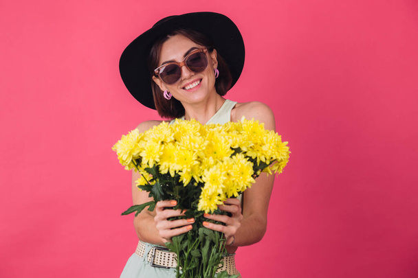 Beautiful stylish woman in hat and sunglasses posing over pink red background holding large bouquet of yellow asters, spring mood, positive emotions isolated space pull to camera - Photo, Image
