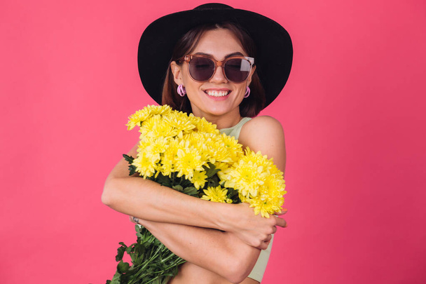 Stylish woman in hat and sunglasses on pink red background hugging large bouquet of yellow asters, spring mood, calm smiling isolated space - Foto, imagen