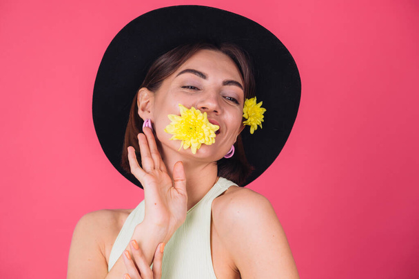 Стильная женщина в шляпе на розово-красном фоне улыбается с двумя желтыми глазами, симпатичная держит один цветок во рту весенним настроением, счастливые эмоции изолированы пространством - Фото, изображение
