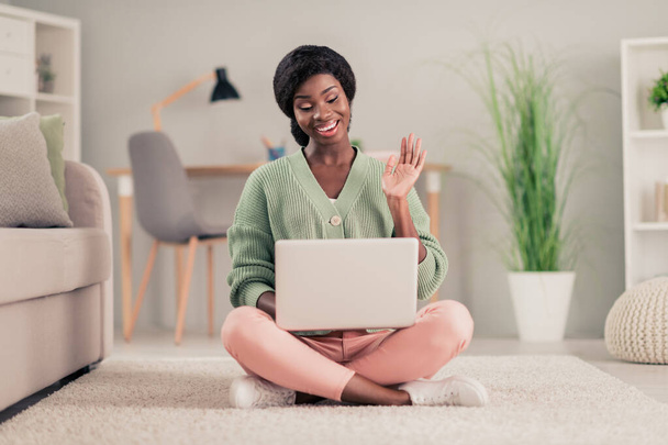 Full length photo of young cheerful afro girl happy positive smile wave hello videocall laptop conference meeting sit floor indoors - 写真・画像