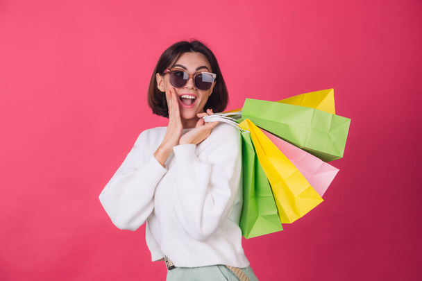 Woman in casual white sweater and sunglasses on pink red background holding shopping bags shocked amazed excited  isolated copy space - Zdjęcie, obraz