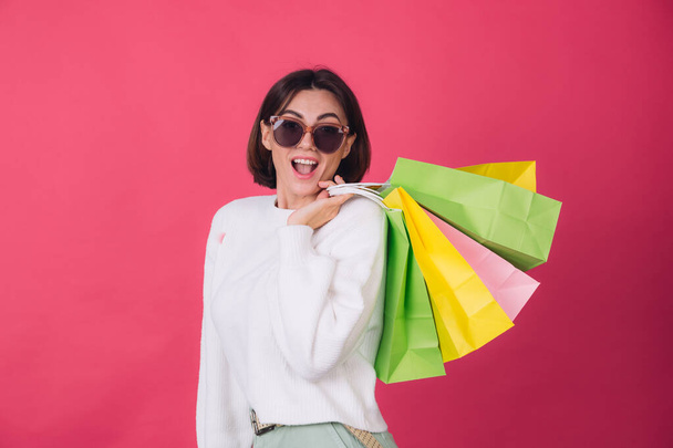 Woman in casual white sweater and sunglasses on pink red background holding shopping bags shocked amazed excited  isolated copy space - Foto, imagen