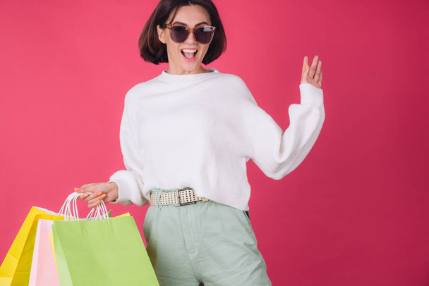 Woman in casual white sweater and sunglasses on pink red background holding shopping bags happy jumping dancing excited isolated copy space - Foto, Bild