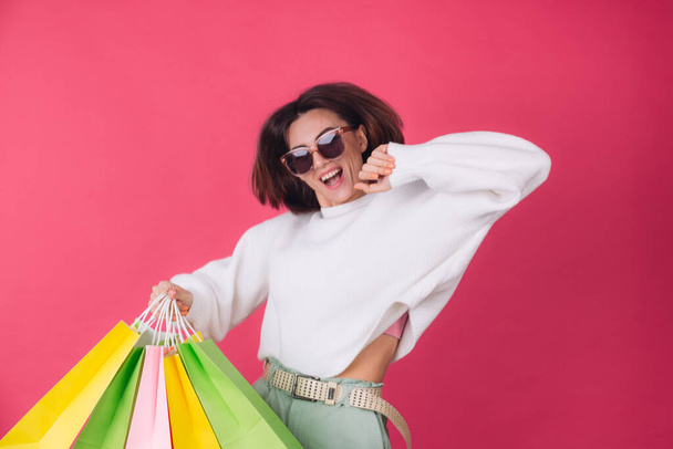 Woman in casual white sweater and sunglasses on pink red background holding shopping bags happy jumping dancing excited isolated copy space - Photo, image