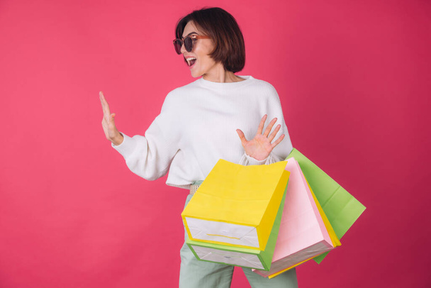 Woman in casual white sweater and sunglasses on pink red background holding shopping bags happy jumping dancing excited isolated copy space - Fotoğraf, Görsel