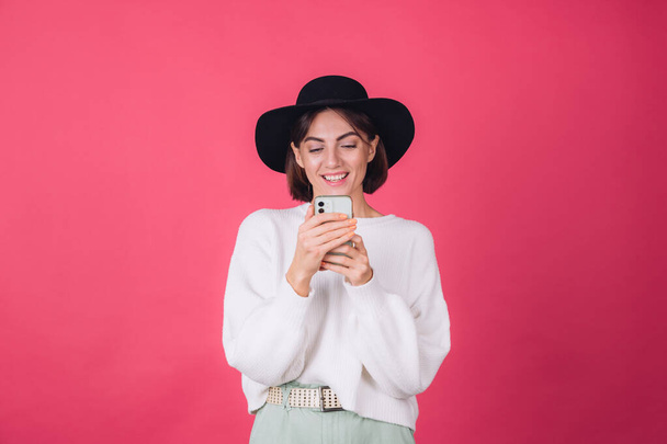 Stilvolle Frau in weißem lässigen Pullover und Hut auf rotem rosa Hintergrund kopieren Raum Lächeln fröhlich halten Mobiltelefon - Foto, Bild