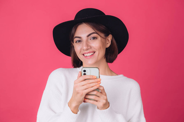 Stylish woman in white casual sweater and hat on red pink background copy space smile cheerful hold  mobile phone - Foto, Bild
