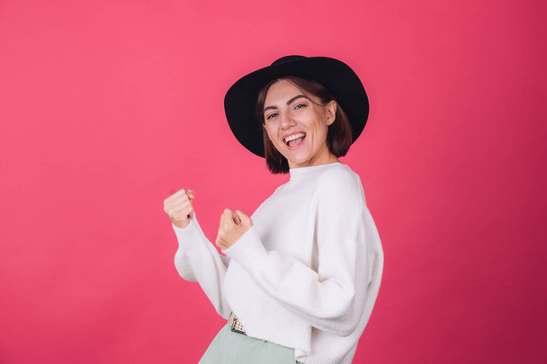 Stylish woman in white casual sweater and hat on red pink background copy space do winner gesture clenching fist celebration - Photo, image
