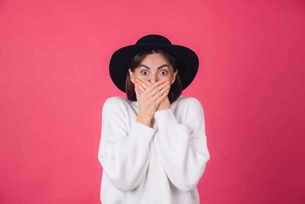 Stylish woman in white casual sweater and hat on red pink background copy space shocked look to camera cover mouth with hands - Fotó, kép