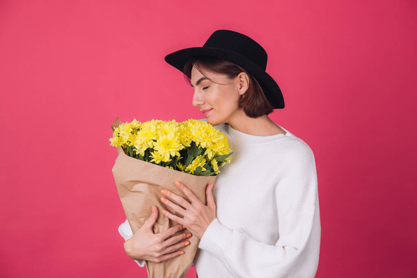 Стильная женщина в шляпе на розовом красном фоне нюхает большой букет желтых цветов, весеннее настроение, счастливые эмоции изолированное пространство с закрытыми глазами - Фото, изображение