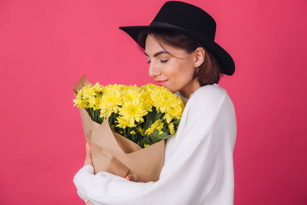 Femme élégante en chapeau sur fond rose rouge odeur renifle grand bouquet de fleurs jaunes, humeur printanière, émotions heureuses espace isolé avec les yeux fermés - Photo, image