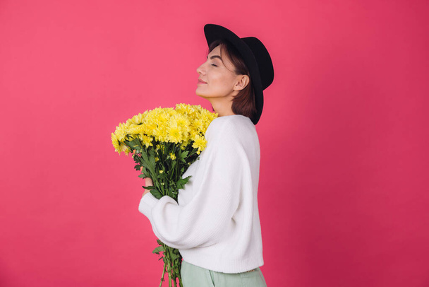 Stijlvolle vrouw in hoed en casual witte trui op roze rode achtergrond houden boeket van gele bloemen, voorjaarsstemming, geïsoleerde ruimte gelukkig glimlach - Foto, afbeelding