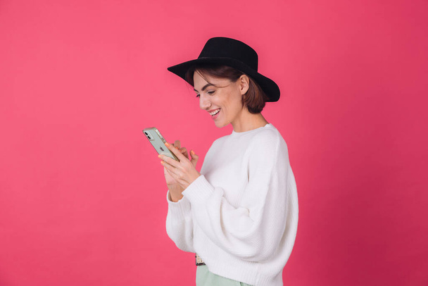 Stilvolle Frau in weißem lässigen Pullover und Hut auf rot rosa Hintergrund Kopierraum Lächeln fröhlich halten Handy Seitenansicht überrascht - Foto, Bild