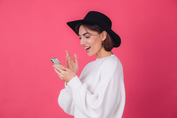 白いカジュアルなセーターと赤いピンクの背景の帽子のスタイリッシュな女性コピースペース笑顔陽気に携帯電話側のビュー驚きを保持 - 写真・画像