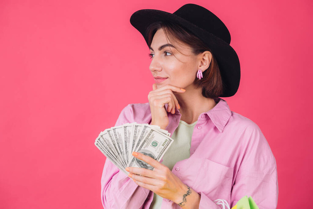 Stilvolle Frau mit Hut auf rosa rotem Hintergrund isoliert Raum nachdenklich halten Fan von 100 Dollar und Einkaufstaschen - Foto, Bild