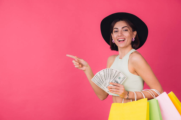 Stilvolle Frau mit Hut auf rosarotem Hintergrund, isoliertes Raumlächeln, aufgeregter Griff eines Fans von 100 Dollar Geld und Einkaufstüten, Zeigefinger nach links - Foto, Bild