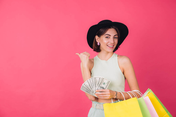 Mujer con estilo en sombrero sobre fondo rosa rojo aislado espacio sonrisa emocionado celebrar ventilador de 100 dólares de dinero y bolsas de compras, dedo pulgar de punta a la izquierda - Foto, imagen