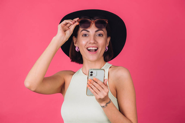 スタイリッシュな女性で帽子とサングラスオンピンク赤の背景隔離されたコピーは驚きの感情を持つ携帯電話を保持 - 写真・画像