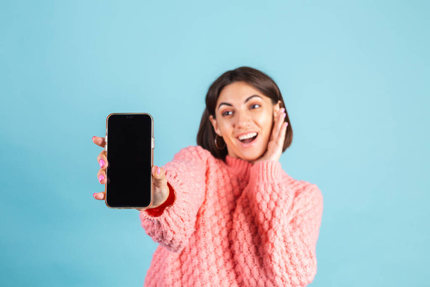 Mladá brunetka v teplém růžovém svetru izolované na modrém pozadí bod na telefonu prázdné černé obrazovce s ohromenou překvapenou tváří - Fotografie, Obrázek