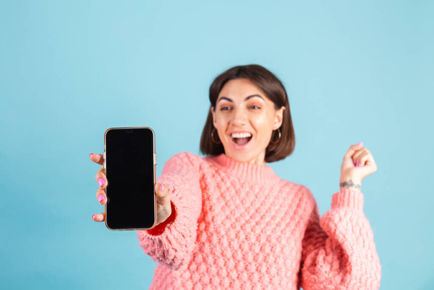 Young brunette in warm pink sweater isolated on blue background point at phone empty black screen with amazed surprised face - Fotó, kép