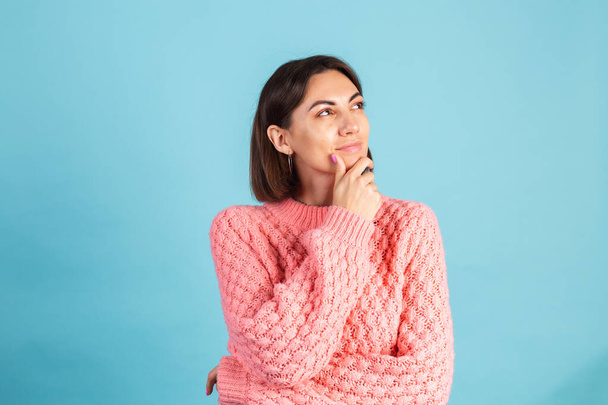 Молодая брюнетка в теплом розовом свитере на голубом фоне задумчиво смотрит в сторону со сладкой улыбкой - Фото, изображение