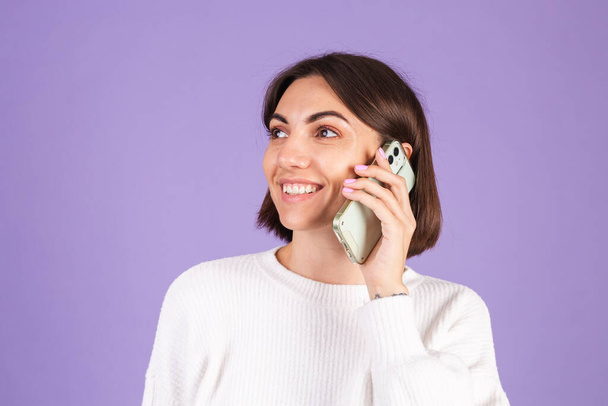 Young brunette in white casual sweater isolated on purple background having conversation with happy mood, talking positive on mobile phone, smiling - 写真・画像