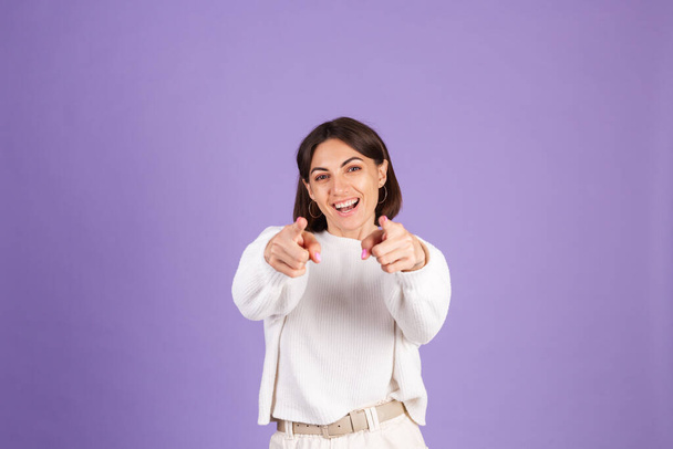 Молодая брюнетка в белом свитере, изолированная на фиолетовом фоне, указывает вам на камеру с счастливым лицом - Фото, изображение