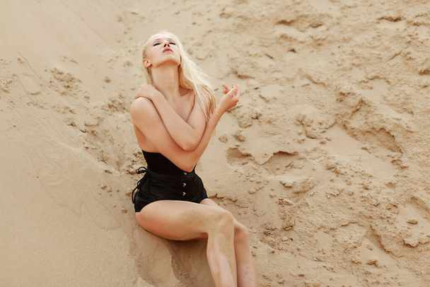 Egy érzéki fiatal nő hosszú szőke hajjal, fekete öltönyben, csukott szemmel fekszik a homokban, kezében egy cigarettával.. - Fotó, kép
