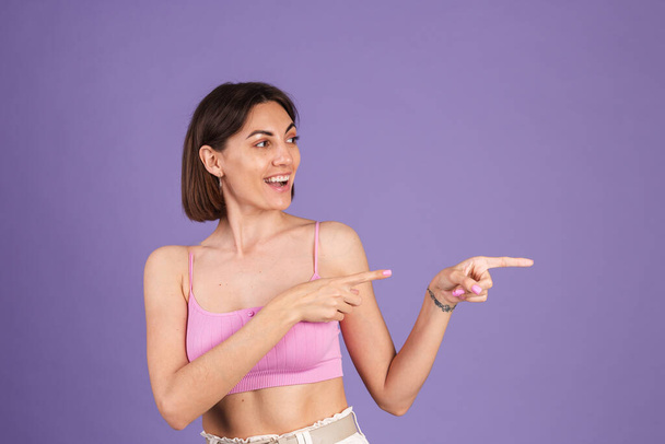 Young brunette in top isolated on purple background fit and pretty happy excited point right with index finger - Photo, Image