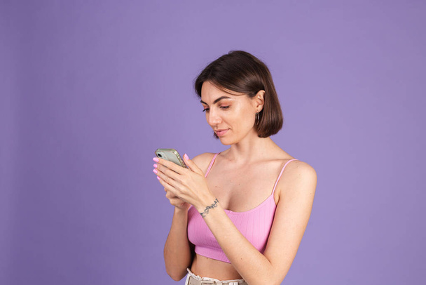 Young brunette in top isolated on purple background holding mobile phone with big smile happy excited face, chatting, having conversation - Foto, Bild