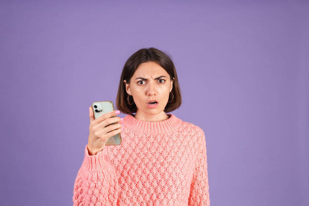 Young brunette in pink sweater isolated on purple background holding phone with shocked disappointed unhappy face - Photo, Image