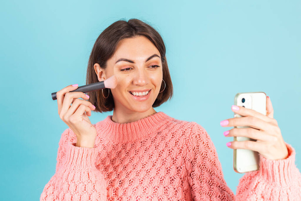 Young brunette in pink sweater isolated on blue background with makeup on face hold blush brush isolated copy space fix makeup holding mobile phone - Fotoğraf, Görsel