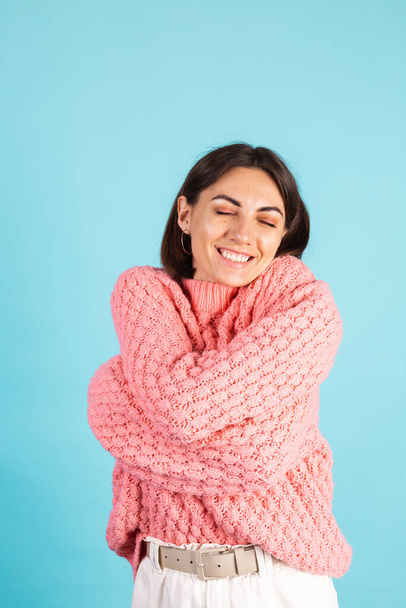 Young brunette in pink sweater isolated on blue background self care hugging herself with smile and closed eyes - Φωτογραφία, εικόνα