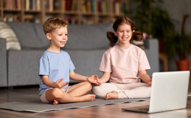 Yoga para niños. Dos niños felices, hermano y hermana en ropa casual sentados en pose de loto en la sala de estar y meditando juntos, mirando a la computadora portátil y sonriendo mientras miran tutorial en línea - Foto, imagen