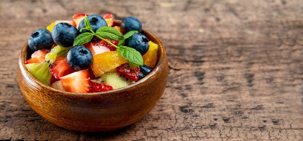 Web banner with fresh summer salad of various fruits and berries. Kiwi, orange, strawberry and blueberry on a wooden bowl decorated with mint on an orange background - Foto, imagen