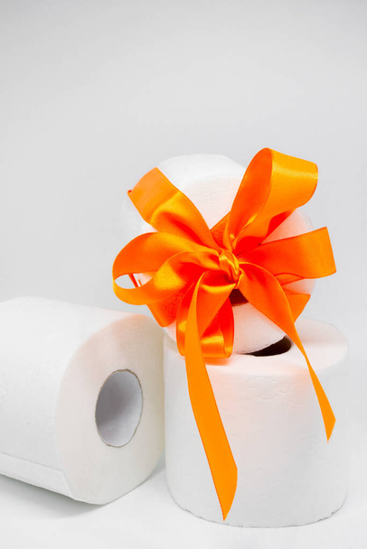 白い穿孔トイレ紙のいくつかのロール。1つのロールは、贈り物として明るいオレンジ色のサテンリボンで結ばれています。垂直写真. - 写真・画像