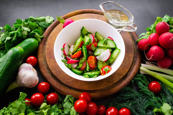 Een stapel verse, rijpe groenten: radijs, tomaten, komkommers, kruiden en knoflook worden rond een ronde houten snijplank gelegd. Een witte kom met groenten gesneden in een salade en olijfolie. - Foto, afbeelding