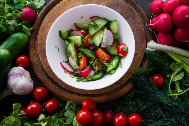 A pile of fresh, ripe vegetables: radishes, tomatoes, cucumbers, herbs, and garlic are arranged around a round wooden chopping board. On it is a white bowl with vegetables cut into a salad. - Foto, Bild