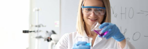 Женщина-ученый в защитных очках разливает химический раствор из колбы в лаборатории - Фото, изображение