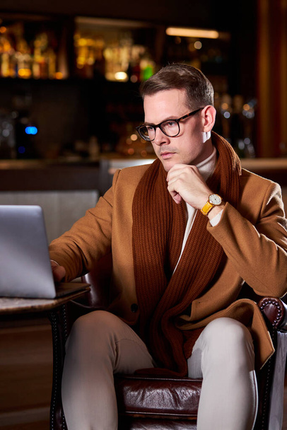 уверенный бизнесмен сидит, думая о деловых сделках и планах, используя ноутбук, в холле гостиницы - Фото, изображение