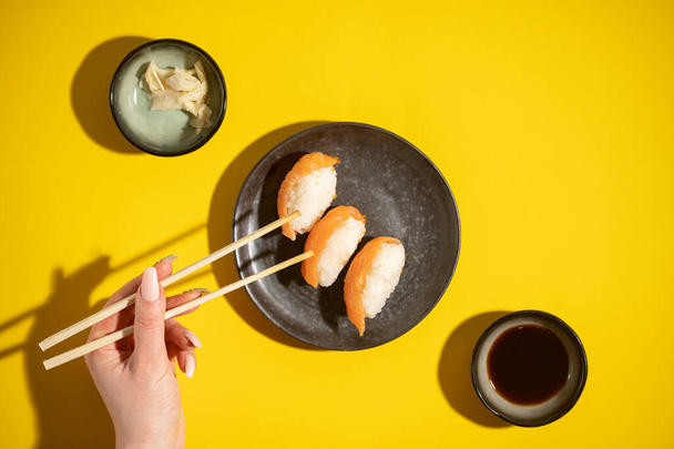 Sushi nigiri con salmone su piatto nero su fondo giallo, con salsa di soja e zenzero e mano femminile con bacchette, spiedino vista dall'alto - Foto, immagini