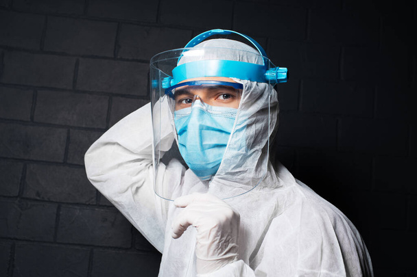 Close-up portrait of young doctor man wearing PPE suit against coronavirus and covid-19. Background of black brick wall. Holding safety face shield with hands. - Photo, Image