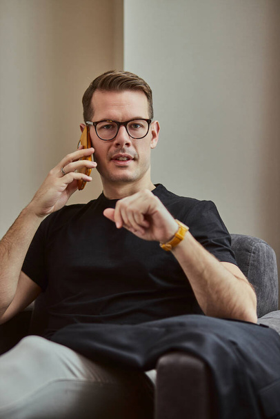 Young businessman talking on smartphone with someone while sitting in hotel room - Photo, Image