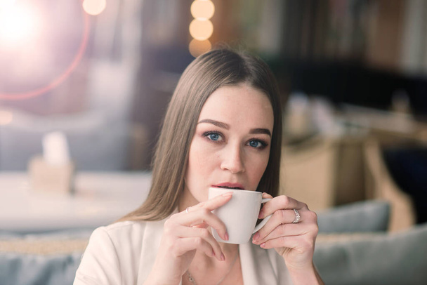 Egy fiatal nő fehér kabátban, aki egy kávézóban dolgozik papírokkal, kávét iszik. - Fotó, kép