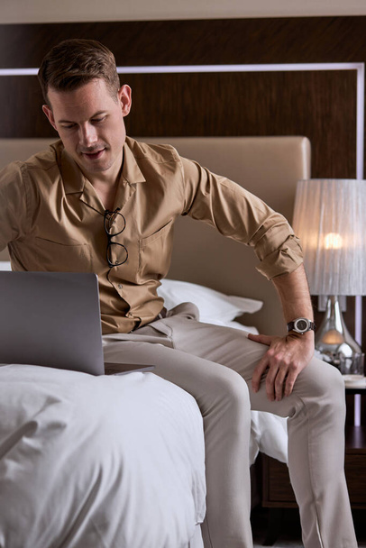 Young handsome guy sit on bed in hotel room, using laptop - Zdjęcie, obraz