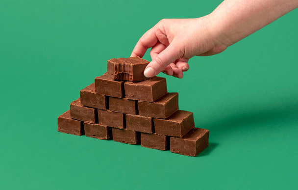 Un colpo minimalista con la mano di una donna che afferra una barretta al cioccolato. Stack di barre fudge isolato su uno sfondo verde. - Foto, immagini