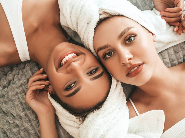 Two young beautiful smiling women in white bathrobes and towels on head.Sexy carefree models lying on bed in posh apartment. They doing beauty treatments at home in towels on heads.After face massage - Fotoğraf, Görsel