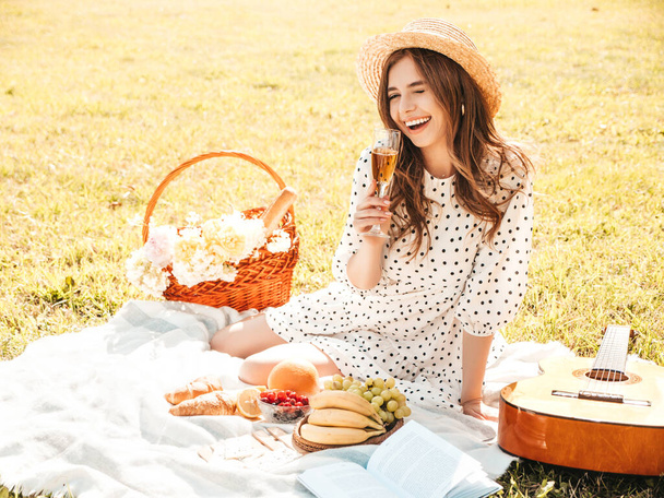 Young beautiful hipster woman in trendy summer sundress and hat. Carefree woman making picnic outside. Positive model sitting on plaid on grass, eating fruits and cheese, drinking champagne - Фото, изображение